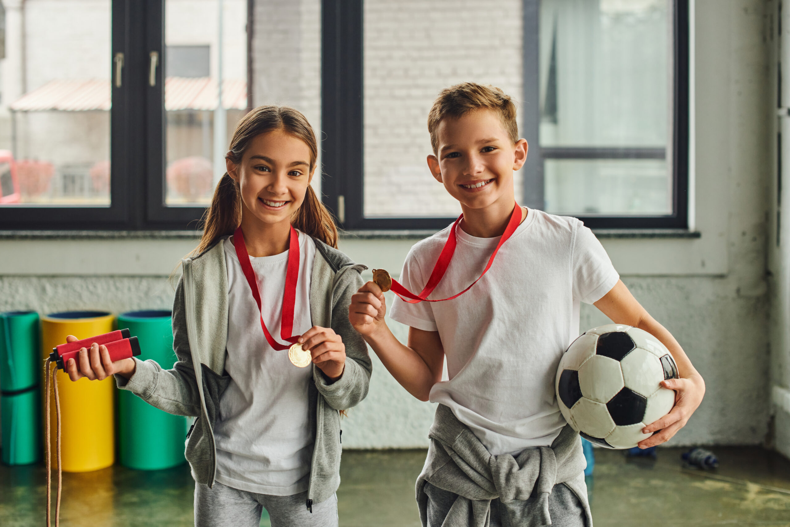 little girl with medal holding jump rope and cute boy posing with soccer ball, smiling at camera