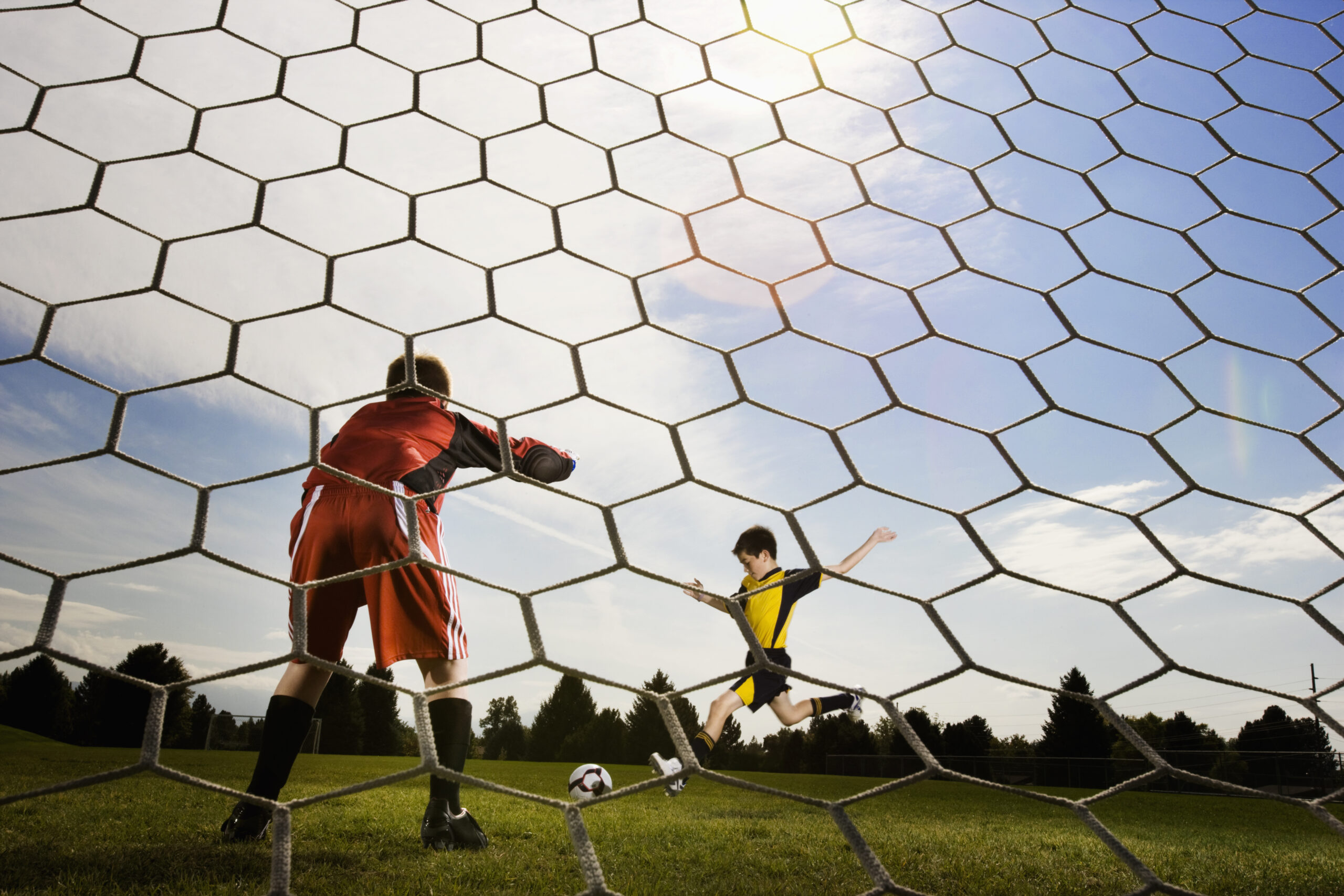 A soccer player taking aim and kicking at the goal, view from behind the goal keeper.